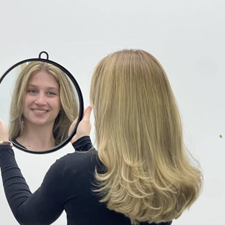 Stranka si v ročnem zrcalu ogleduje svetel blond balayage s stopnjevanim krojem